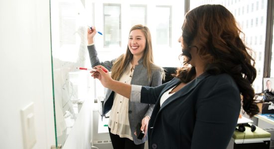 Two professional women discussing ideas on a whiteboard in a modern office setting.