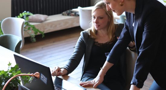 Two professional women discuss a project using a laptop in a modern office environment.